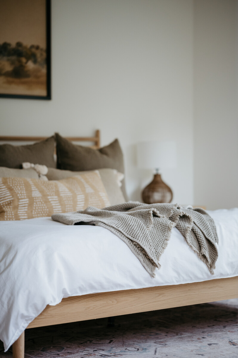 Neutral bedroom with a wood bed frame, white linens, patterned pillows, and a waffle-weave throw, part of Jenna Kutcher's evening wind-down routine designed to calm the nervous system before bed.
